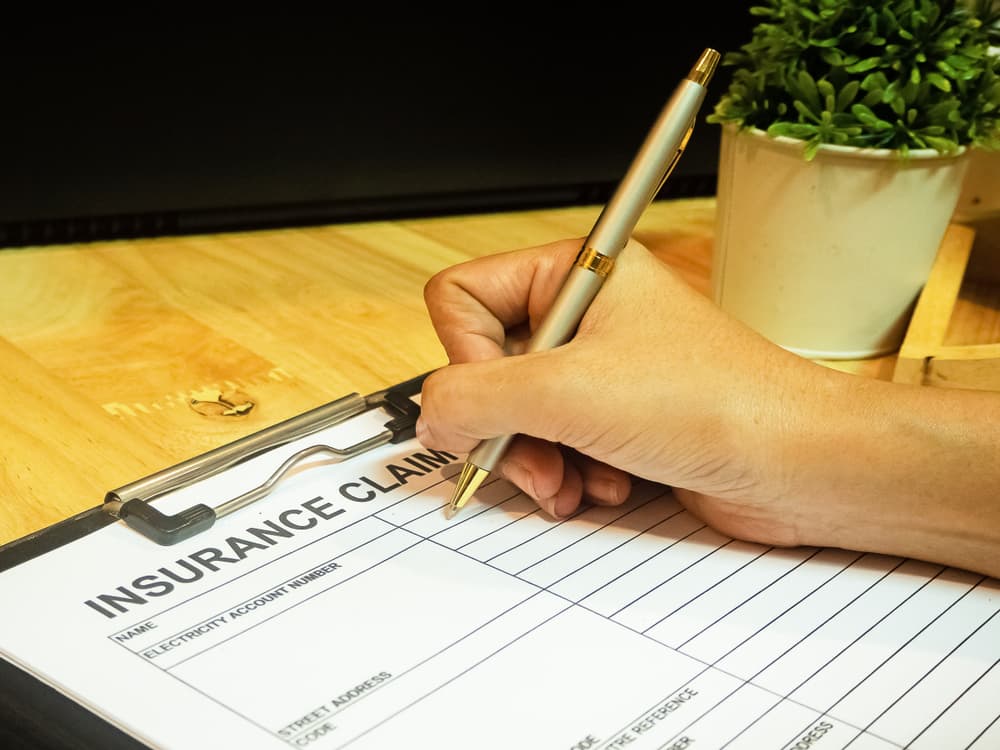 A human hand uses a pen to fill out information on an insurance claim form.