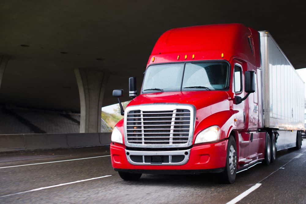 A modern big rig semi truck with a high cabin for improved aerodynamics moves under a bridge, transporting a dry van semi trailer for long-haul freight.






